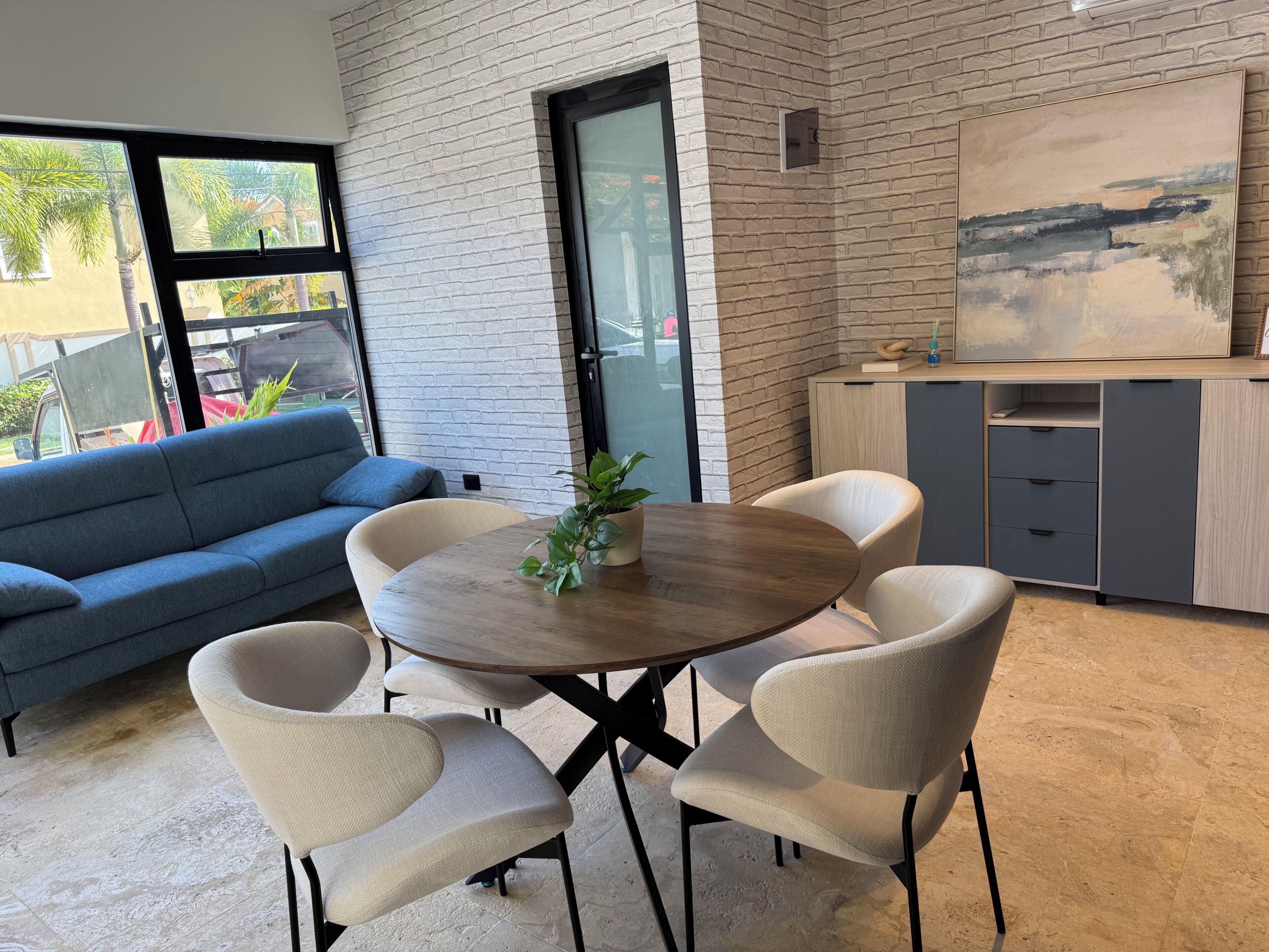 A modern living room with a round wooden table, white cushioned chairs, a blue sofa, and a grey cabinet with a painting.