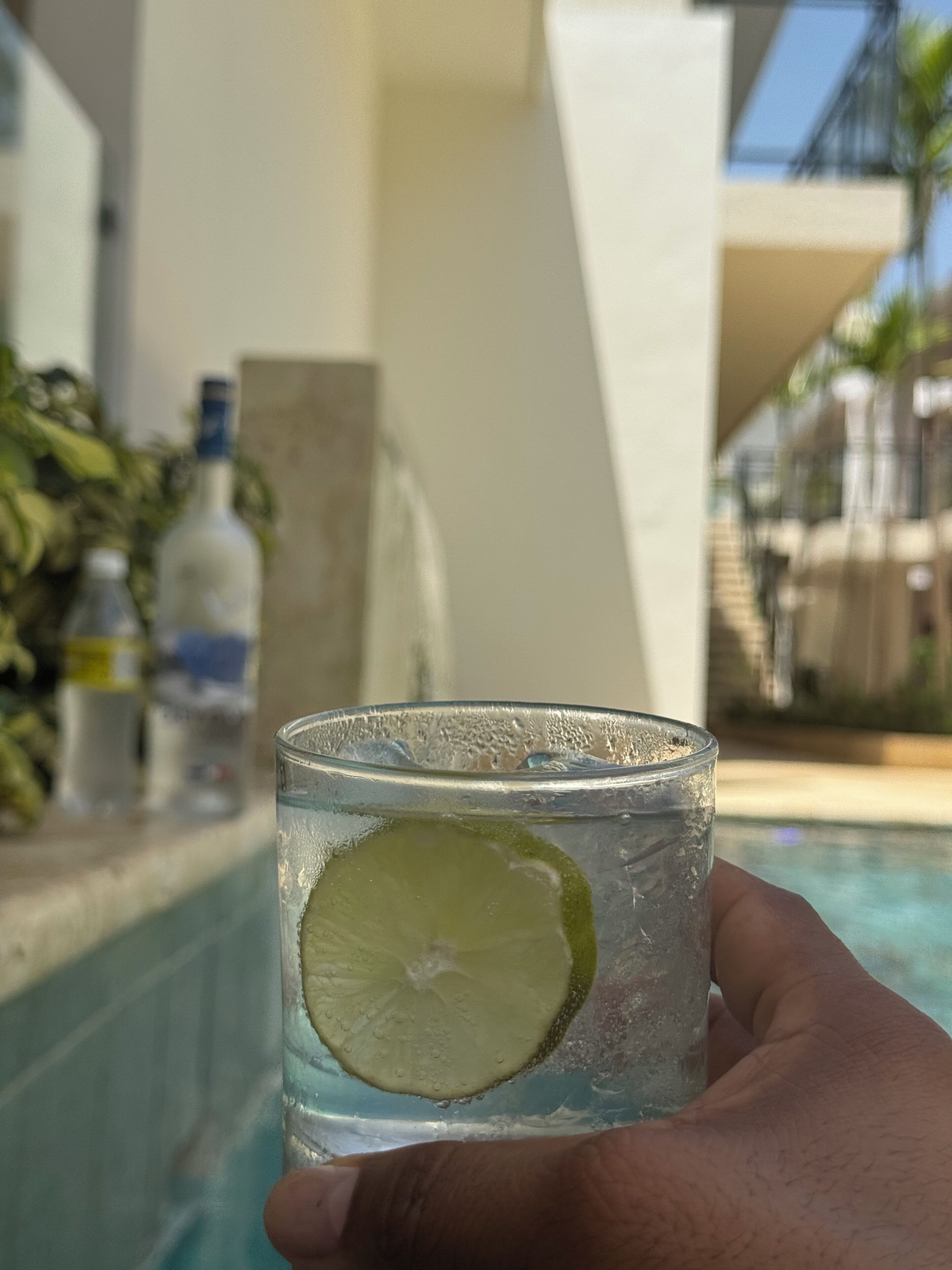 A hand holding a glass with a lime slice and ice, near a pool with a clear view of a building and some plants in the background.
