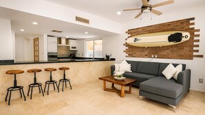 Un salon moderne avec un canapé gris, une table basse et trois tabourets de bar en bois. Une planche de surf est accrochée au mur, au-dessus du canapé.