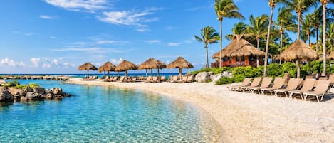 A beach resort with clear turquoise waters, lounge chairs, and thatched umbrellas.