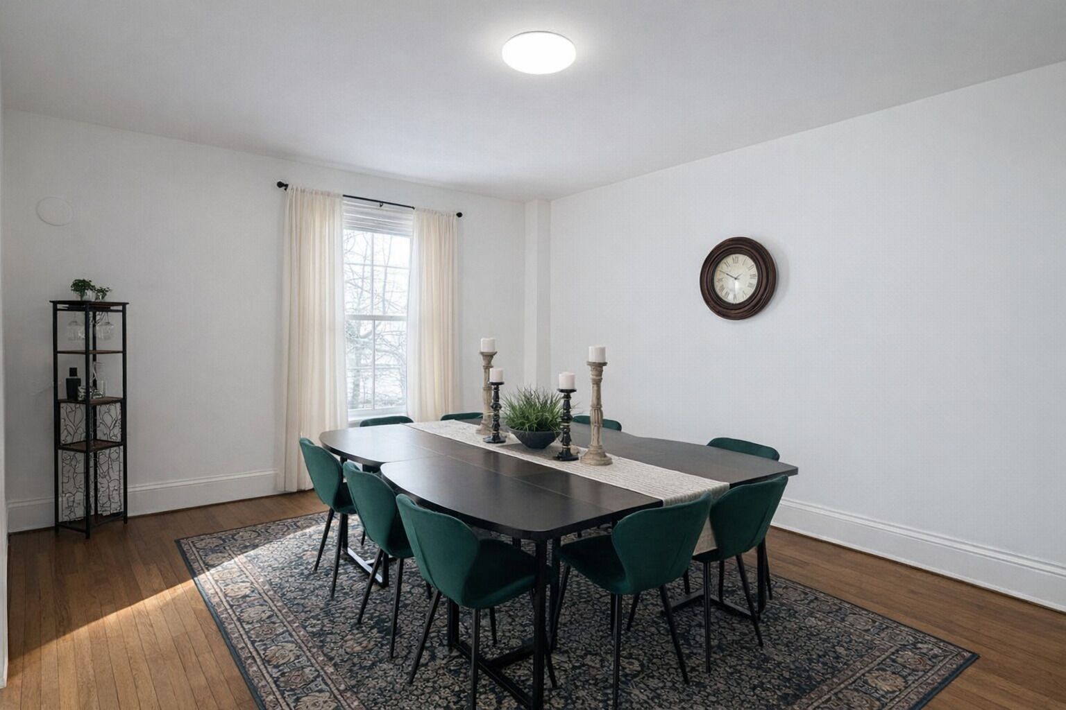 A dining room with a dark wooden table, green chairs, a patterned rug, a tall candle holder, and a clock on the wall.