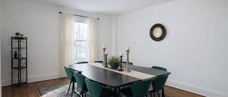 A dining room with a dark wooden table, green chairs, a patterned rug, a tall candle holder, and a clock on the wall.