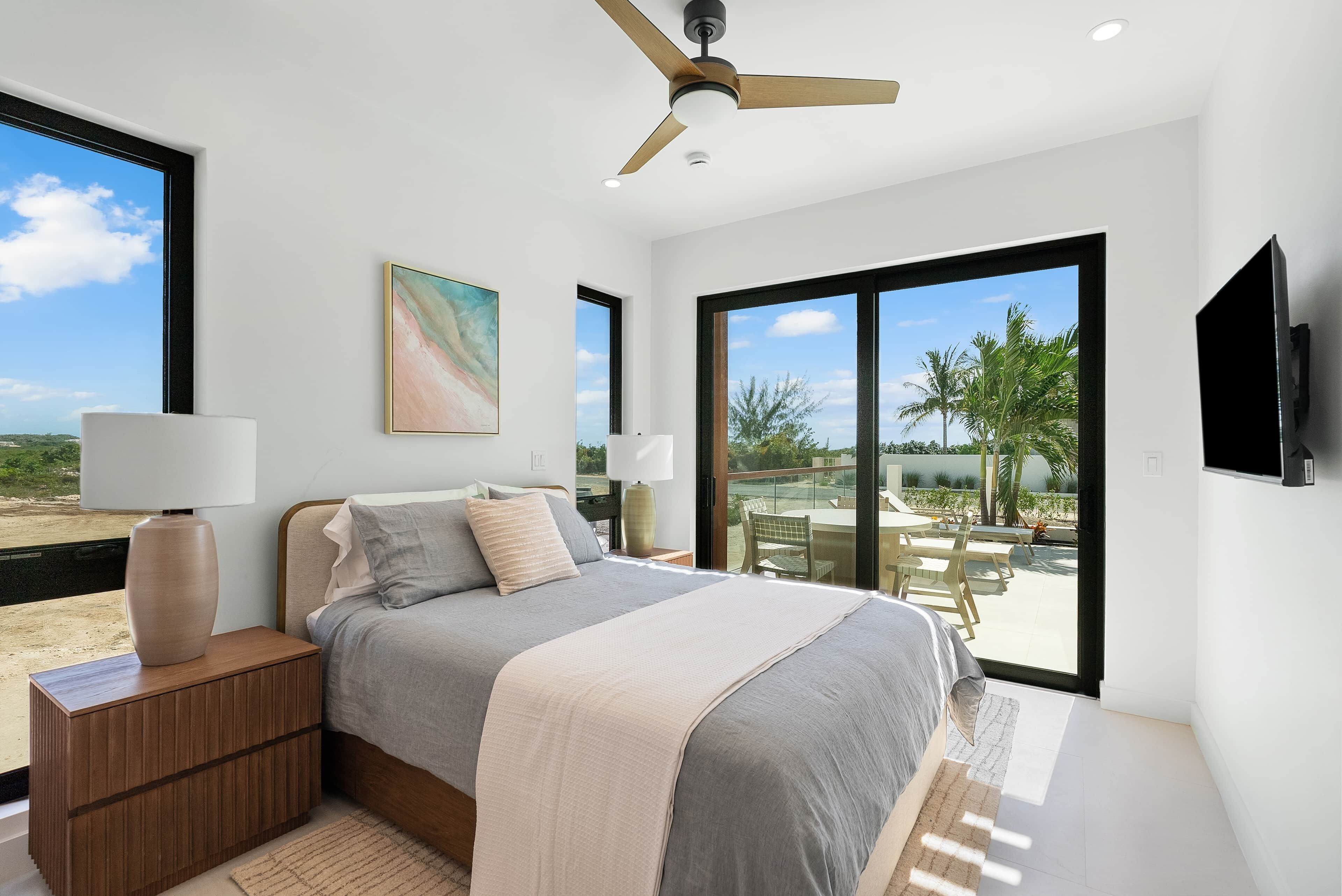 A modern bedroom with a large bed, a ceiling fan, and a view of palm trees through the sliding glass door.