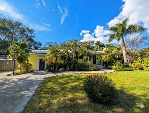A yellow house with a gravel driveway, surrounded by palm trees and lush greenery. - Coastal Canopy, Private Pool: 15 Min to Beach (Bradenton)