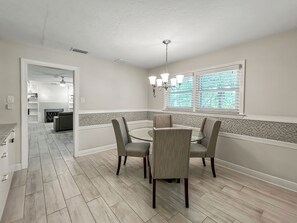 A dining area with a glass table, four chairs, a chandelier, and a view through a window with blinds. - Coastal Canopy, Private Pool: 15 Min to Beach (Bradenton)