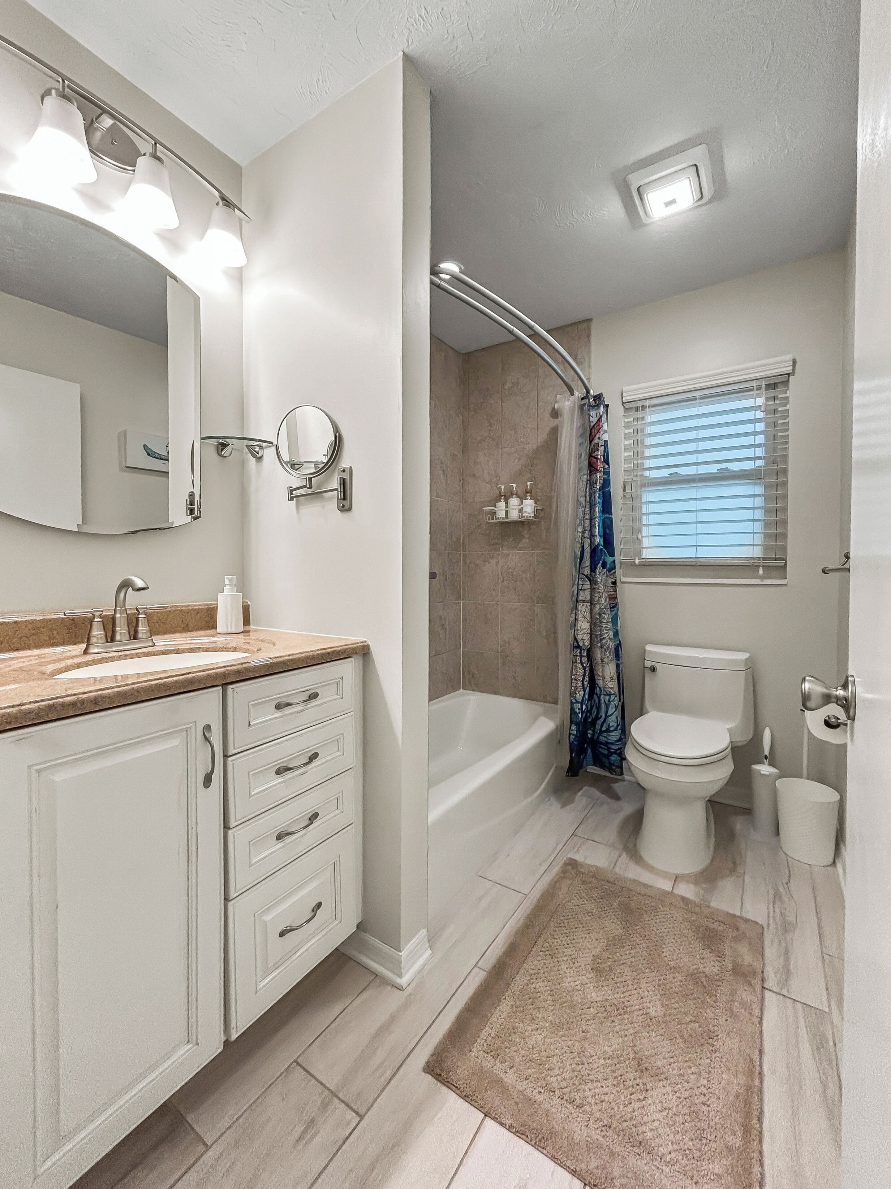 A bathroom with a sink, mirror, vanity, shower, toilet, and a window with blinds.