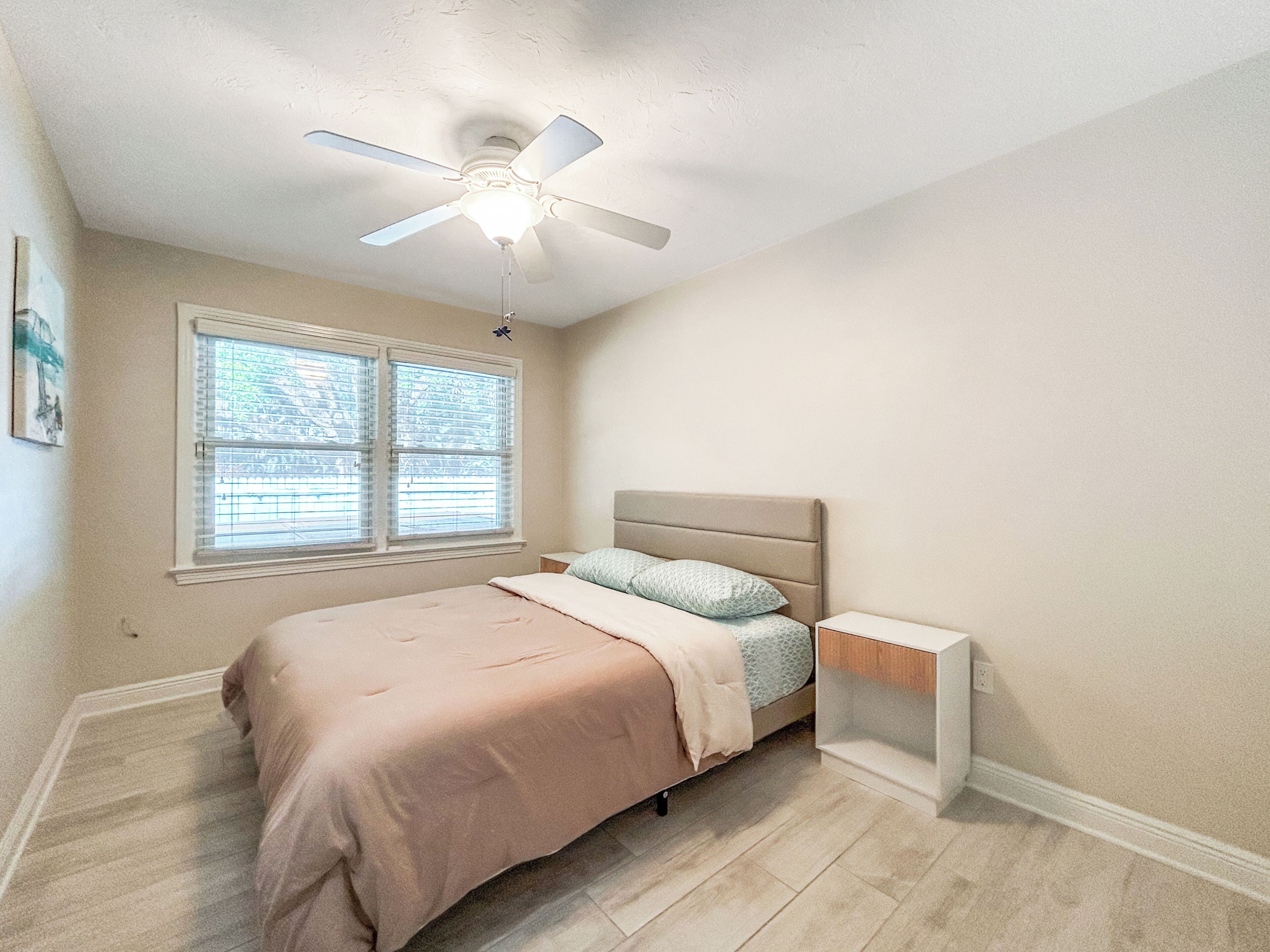 Une chambre à coucher comprenant un lit, une table de chevet, un ventilateur de plafond et une fenêtre avec des stores.
