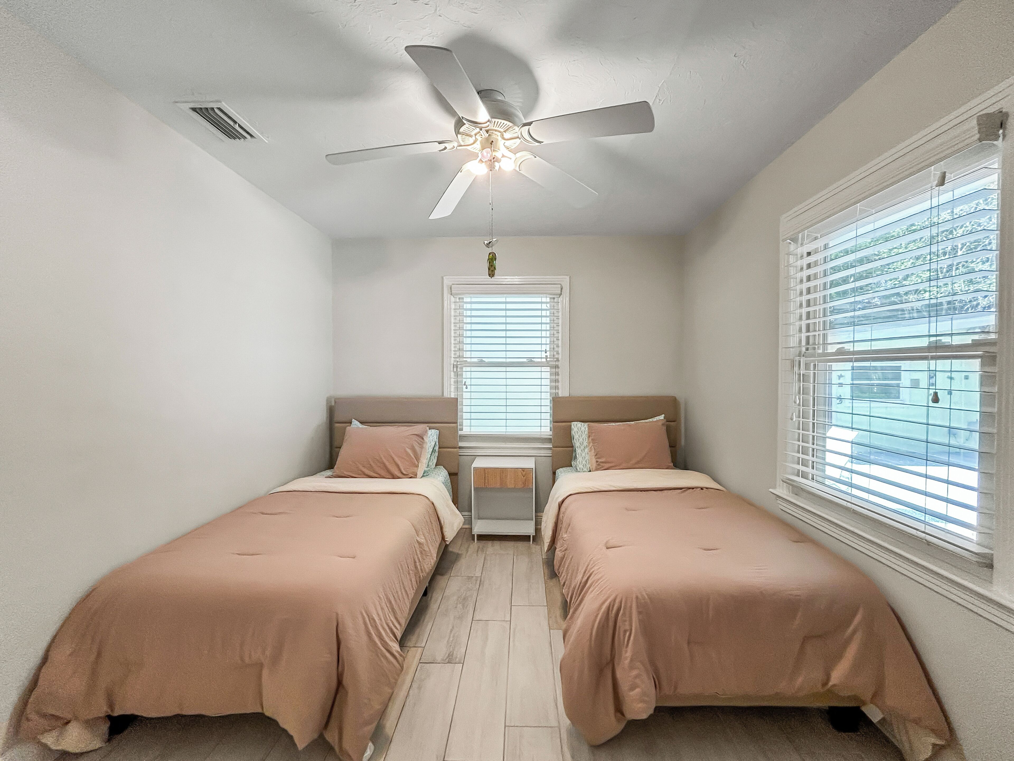 Une chambre avec deux lits, un ventilateur de plafond, une fenêtre avec des stores et une table de chevet.