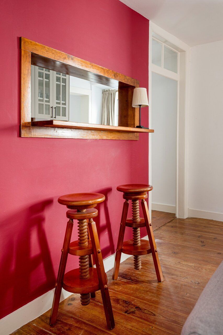 Ein Zimmer mit roter Wand, einem spiegelnden Rahmen aus Holz, zwei Holzschemeln und einer weißen Lampe auf einem Regal.