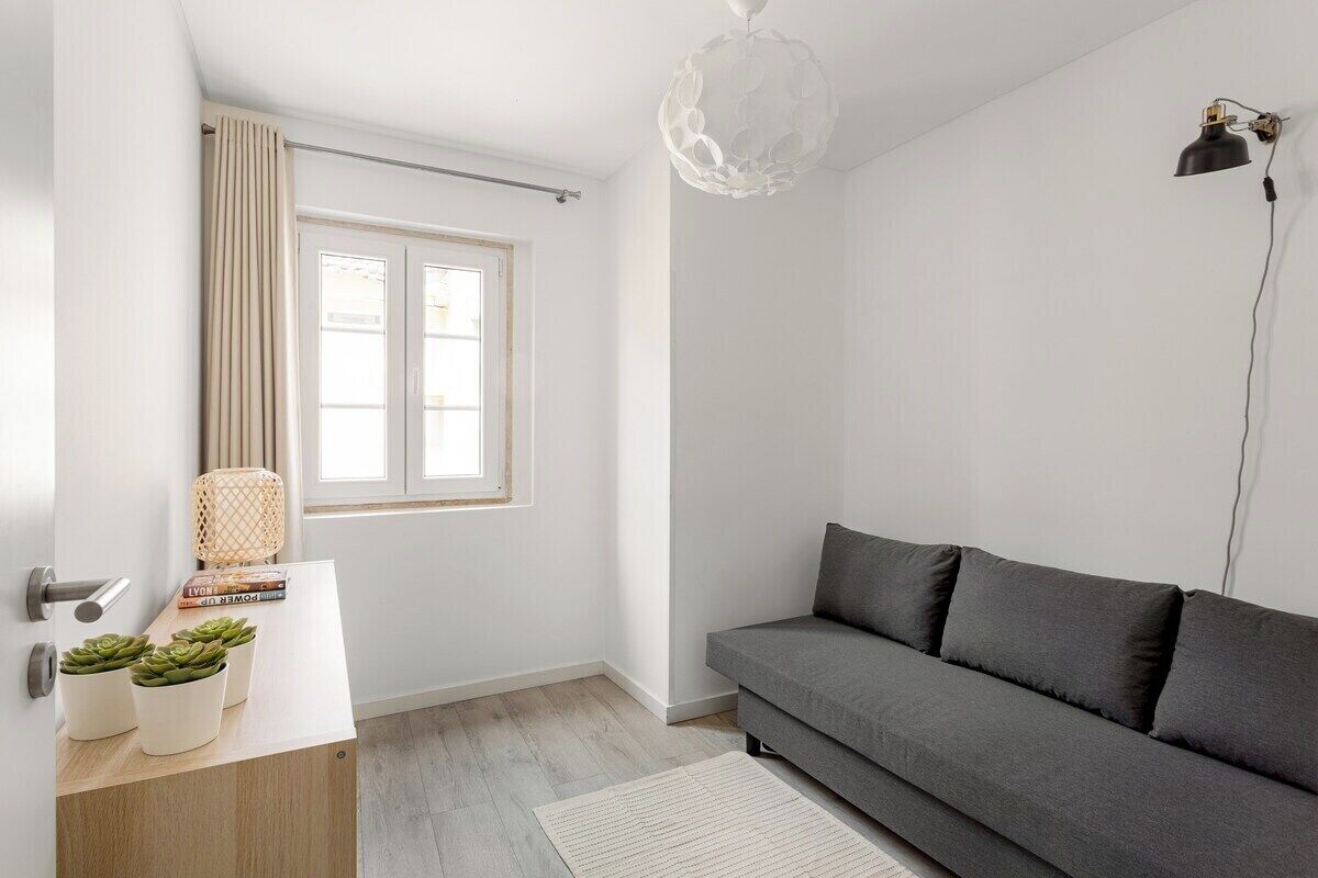 Una sala moderna con un sofá gris, suelo de madera, pared blanca, ventana con cortinas, lámpara colgante y una mesita con libros y plantas.