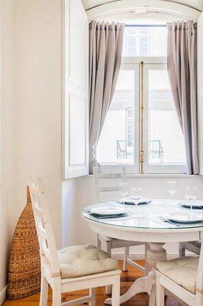 A dining area with a round table, white chairs, and a window with curtains. - Baixa-Chiado Getaway (Lisbon)