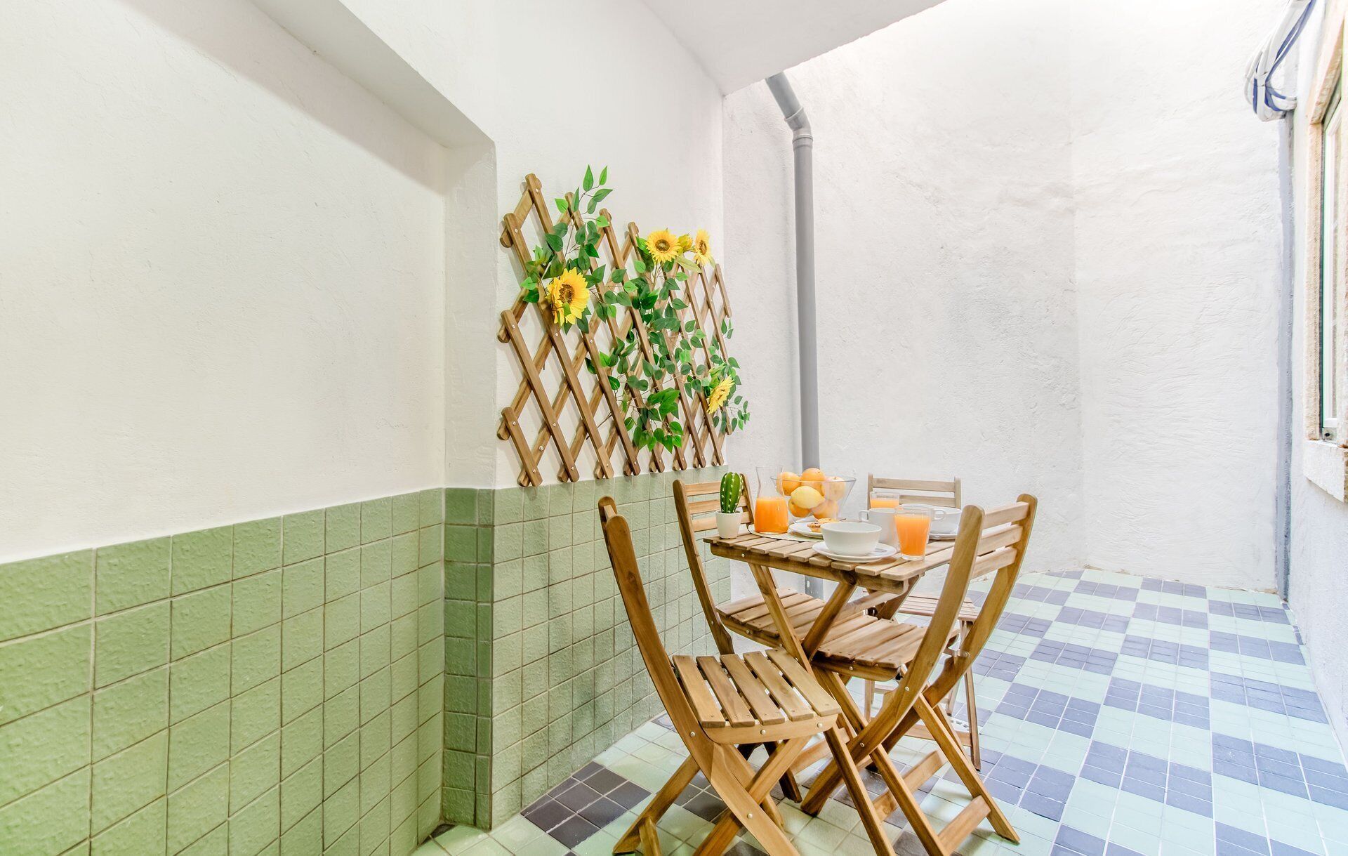 A small, enclosed dining area with a checkered floor, wooden chairs, a table with a glass of orange juice, and a decorative wall with sunflowers and a wooden lattice.