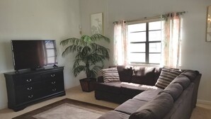 Ein Wohnzimmer mit einer Couch, einem Fernseher auf einem Schrank, einer großen Pflanze und einem Fenster mit Vorhängen.