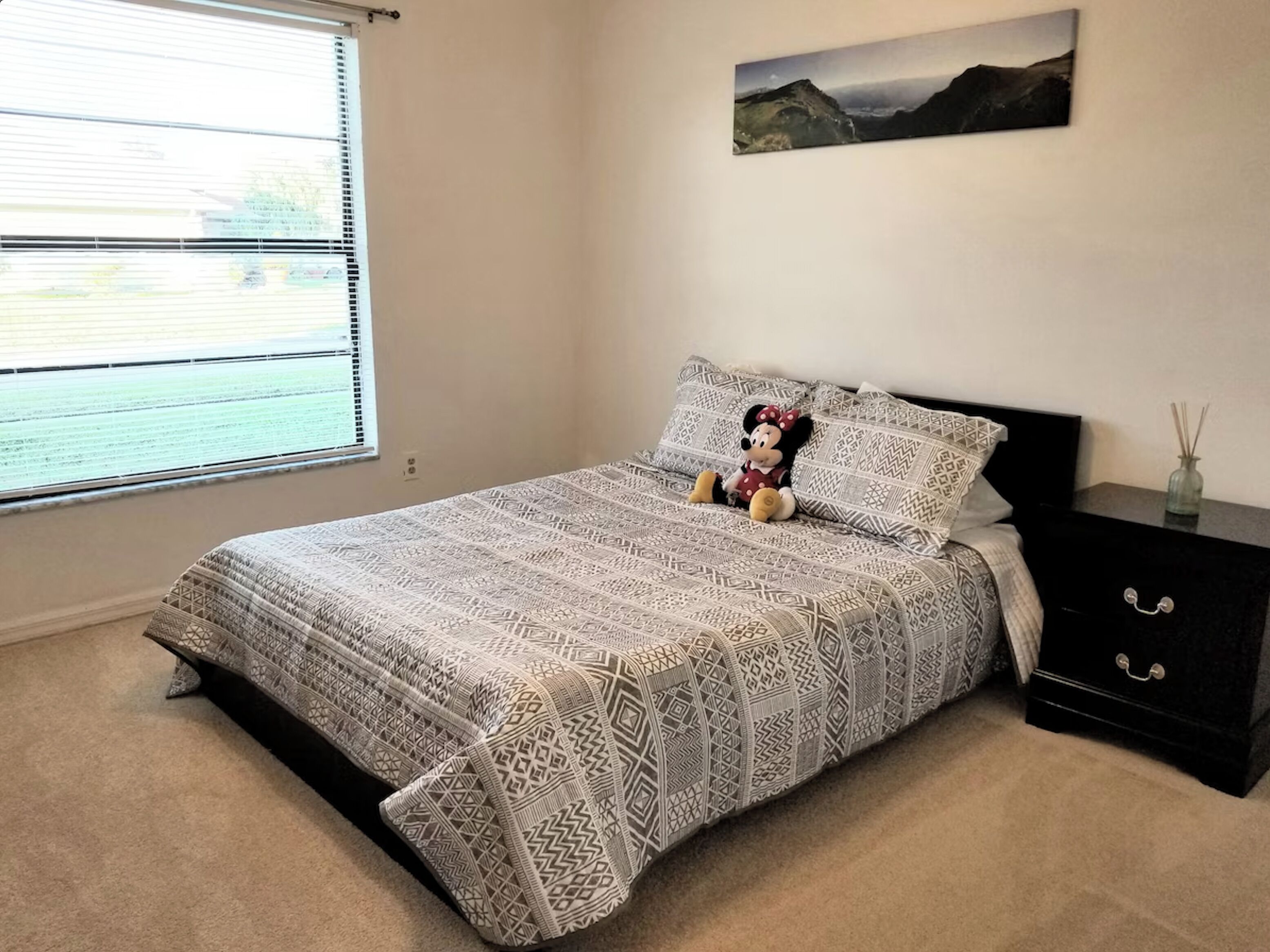 Un dormitorio con una colcha estampada, un peluche de Mickey Mouse, una mesita de noche con cajones, una ventana con persianas y un cuadro de paisaje en la pared.