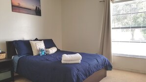 Ein Schlafzimmer mit Bett, Kissen, Decke, einem kleinen Tisch mit Korb und einem Fenster mit Vorhängen.