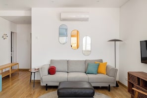 Ein modernes Wohnzimmer mit grauem Sofa, Hocker in Schwarz, einem Holztisch und einer Stehlampe.