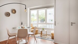 Ein Zimmer mit einem Esstisch, Stühlen, einem kleinen runden Tisch mit einer Pflanze und einem großen Fenster mit Blick nach draußen.