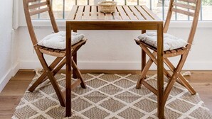 Ein Essbereich mit Holztisch und Stühlen, einem gewebten Teppich und Blick durch große Fenster mit Jalousien.