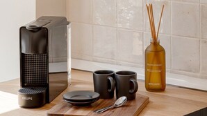 Eine Küchenzeile mit einer Kaffeemaschine, zwei Tassen, einem Löffel, einer Flasche ätherischem Öl und einem Holztisch.