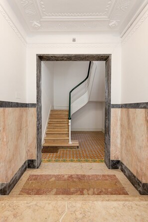 Eine Marmortreppe mit einem gemusterten Boden führt zu einem Raum mit weißen Wänden und einer Decke mit dekorativer Stuckverzierung.