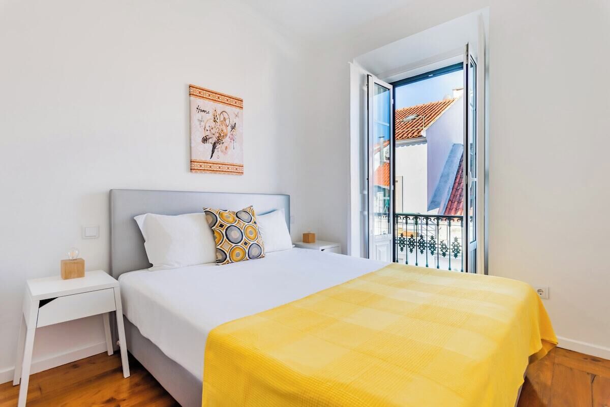 Ein Schlafzimmer mit einem Bett, einem Nachttisch, einem Fenster mit Blick auf Gebäude und einer Wand mit einem gerahmten Bild.