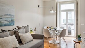 A modern living room with a grey sofa, a dining table with chairs, a pendant light, and a large window with a view of the city. - Trendy Flat in the iconic Chiado (Lisbon)