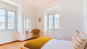 Un dormitorio con una cama, dos ventanas, una silla y una obra de arte colgada en la pared.