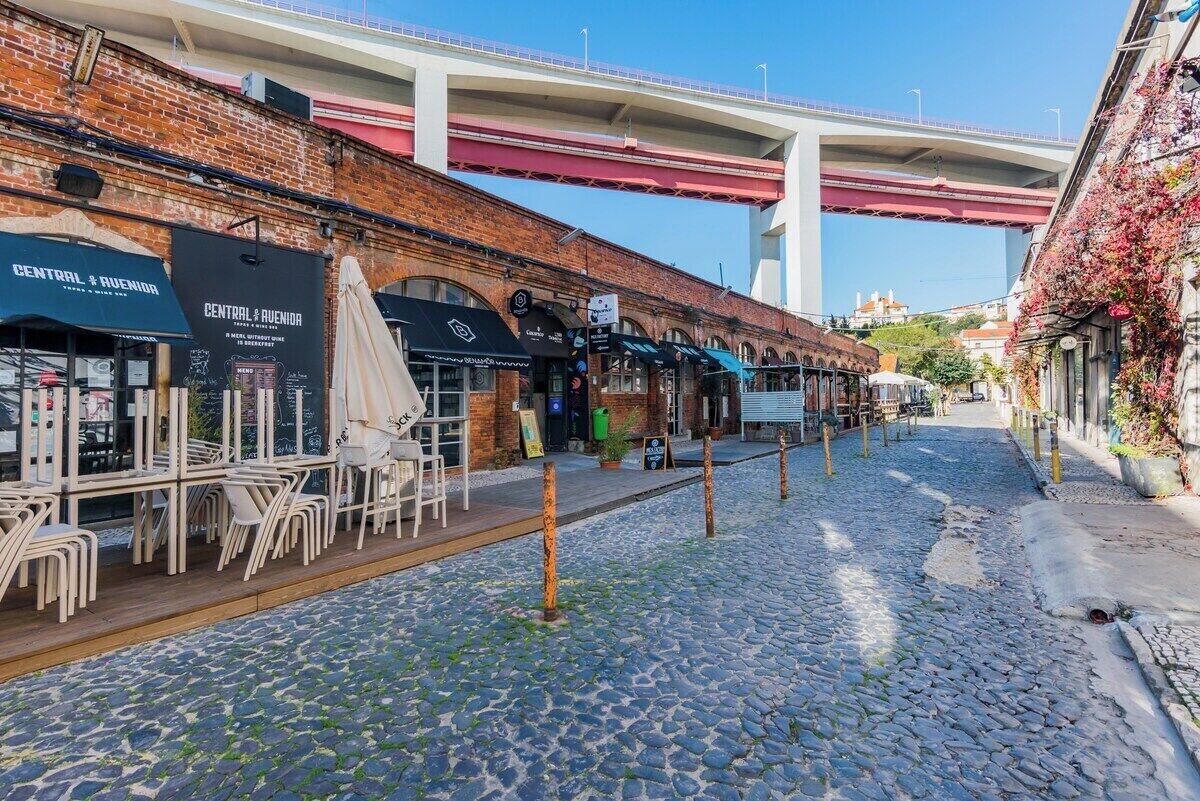 A cobblestone street with outdoor seating, a brick building, and a bridge in the background.