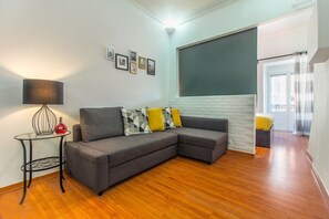 Ein Wohnzimmer mit einem grauen Ecksofa, einem kleinen runden Tisch mit Lampe und einer weißen Ziegelwand.