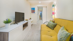 Un espai de viure reduït amb un sofà, una televisió sobre un moble blanc i una planta petita.