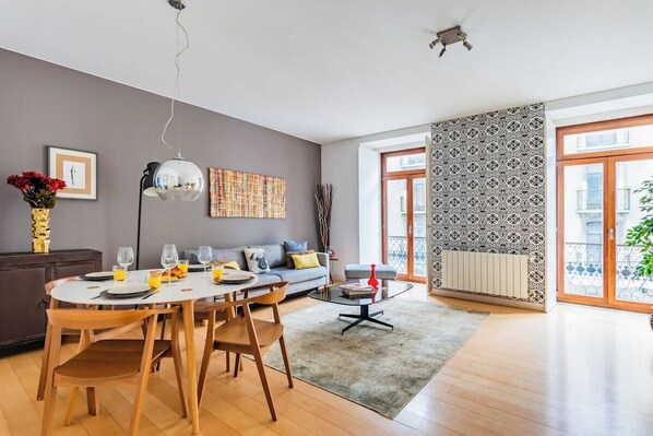 A modern living room with a dining area, a sofa, a coffee table, and a large window with patterned curtains. - Chiado Mercy II with AC (Lisbon)