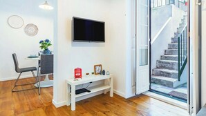 En moderne stue med et fladskærms-tv, et spiseområde med bord og stole, et lille hvidt konsolbord med pynteting, og en trappe, der fører til et udendørs område.