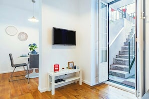 Ein modernes Wohnzimmer mit Flachbildfernseher, einem Essbereich mit Tisch und Stühlen, einem kleinen weißen Konsolentisch mit Dekoration und einer Treppe, die zu einem Außenbereich führt.