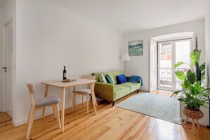 Ein Zimmer mit Holzboden, einem grünen Sofa mit blauen Kissen, einem kleinen Tisch mit Flasche und Gläsern sowie einer Zimmerpflanze.