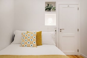 Ein Bett mit weißen und gemusterten Kissen, ein kleines Regal mit einer Pflanze und eine weiße Tür.