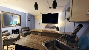 A compact hotel room with a kitchenette, a bed with pillows, a TV, and a ceiling fan. - Quaint 1-bedroom island condo in Corpus Christi with all the comforts of home. (Corpus Christi)