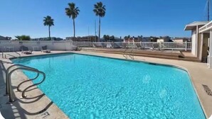 A swimming pool with clear blue water, surrounded by a white fence, palm trees, and a building with a balcony. - Quaint 1-bedroom island condo in Corpus Christi with all the comforts of home. (Corpus Christi)