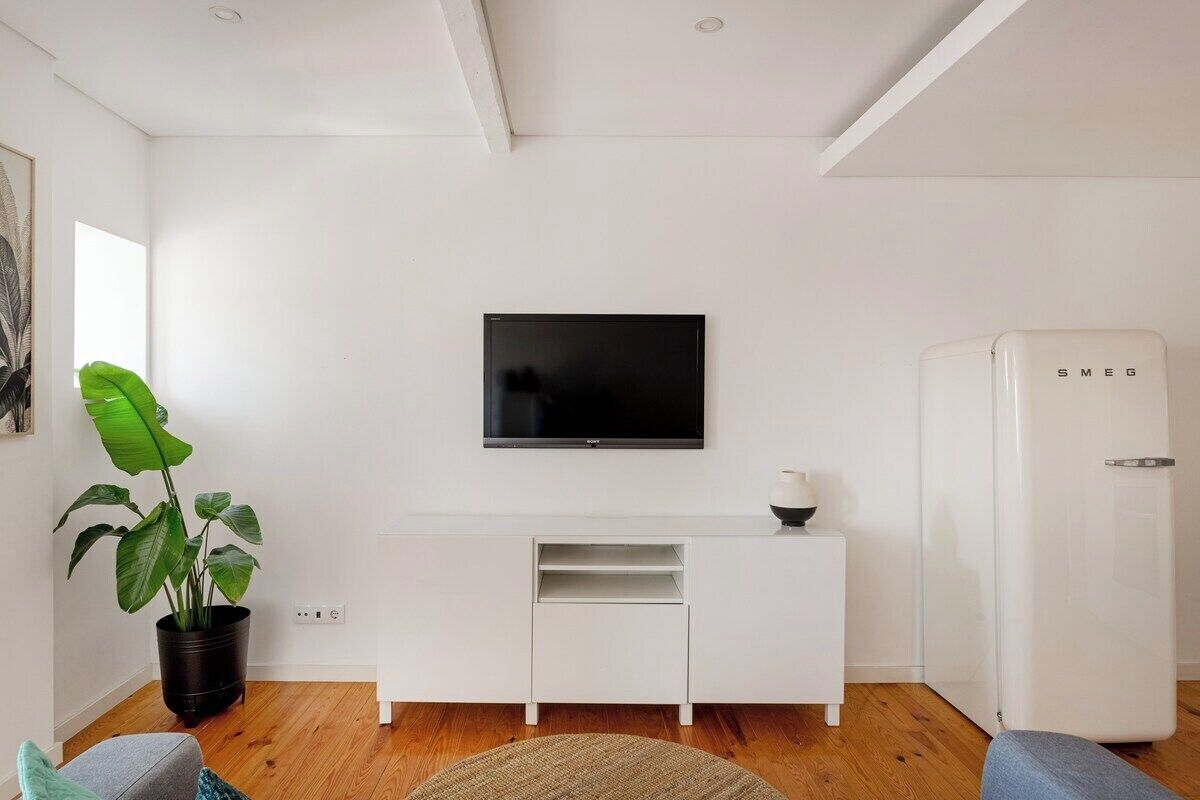 Ein modernes Wohnzimmer mit einem an der Wand befestigten Flachbildfernseher, einem weißen Schrank darunter, einer Topfpflanze, einem Kühlschrank und Holzdielen.