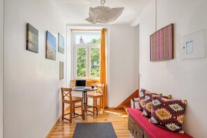 Ein Zimmer mit Schreibtisch, Stühlen, einem Laptop, einem Bett mit gemusterten Kissen und Bildern an den Wänden.