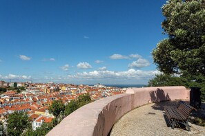 Eine gewölbte Steinmauer mit einer Bank, mit Blick auf eine Stadtlandschaft mit roten Dächern und blauem Himmel.