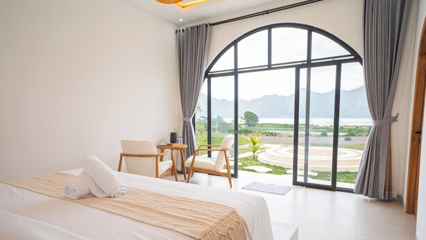 A bedroom with a large bed, two chairs, a small table, and a view of a lake and mountains through a large arched window. - Lazura Serenite (Kintamani)