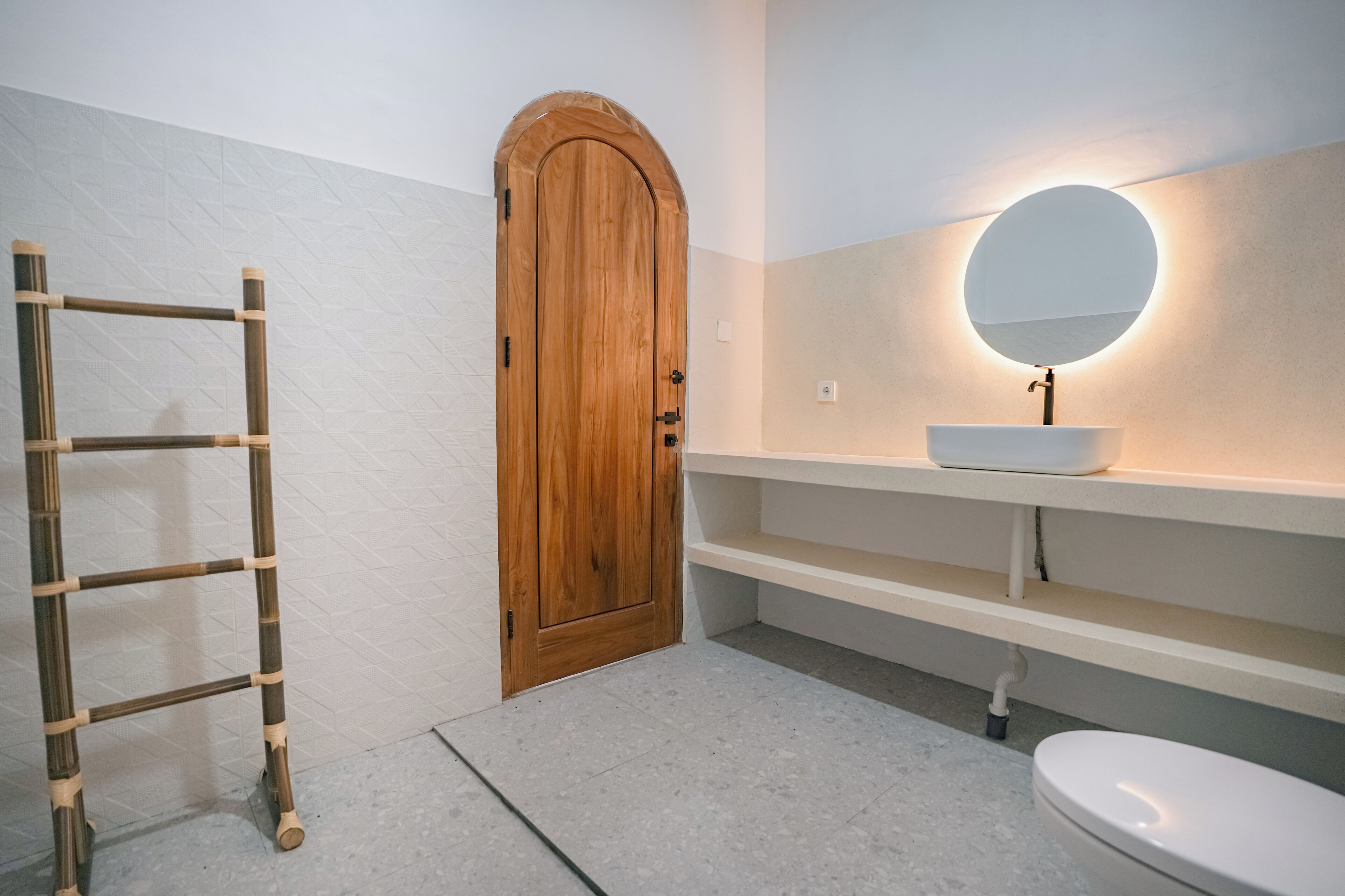 A bathroom with a wooden door, a white sink with a round mirror, and a bamboo ladder.