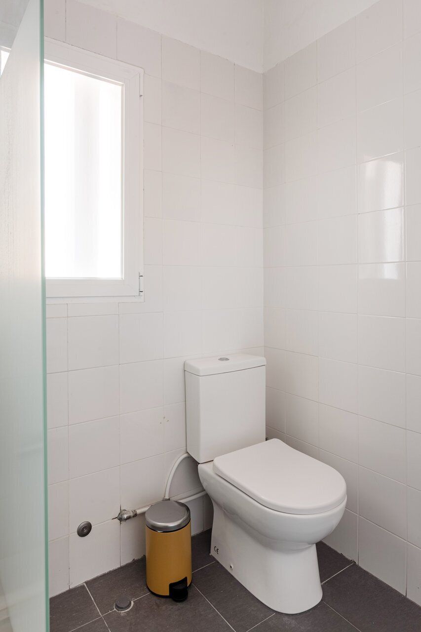 Een badkamer met witte betegelde wanden, een wit toilet, een glazen douchecabine en een kleine prullenbak.