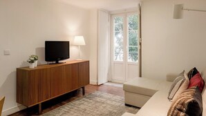 Una sala de estar con un televisor sobre un mueble de madera, un sofá con cojines, una lámpara y una ventana con vistas a árboles.