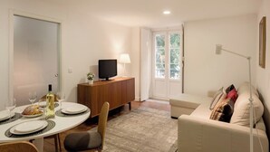 Un comedor con una mesa lista para dos personas, un televisor sobre un mueble de madera y un sofá al fondo.