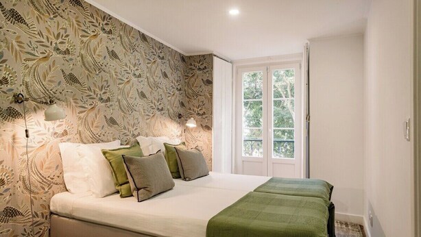 Un dormitorio con cama, almohadas, una manta verde, una ventana y un papel tapiz estampado.