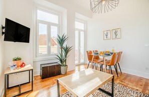 Ein modernes Wohnzimmer mit Holzboden, einem Esstisch mit Stühlen, einem an der Wand montierten Fernseher und einem großen Fenster.