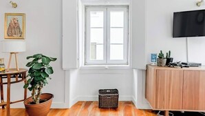 Ett rum med ett stort fönster, en krukväxt, en korg av rotting, ett träskåp och en tv.