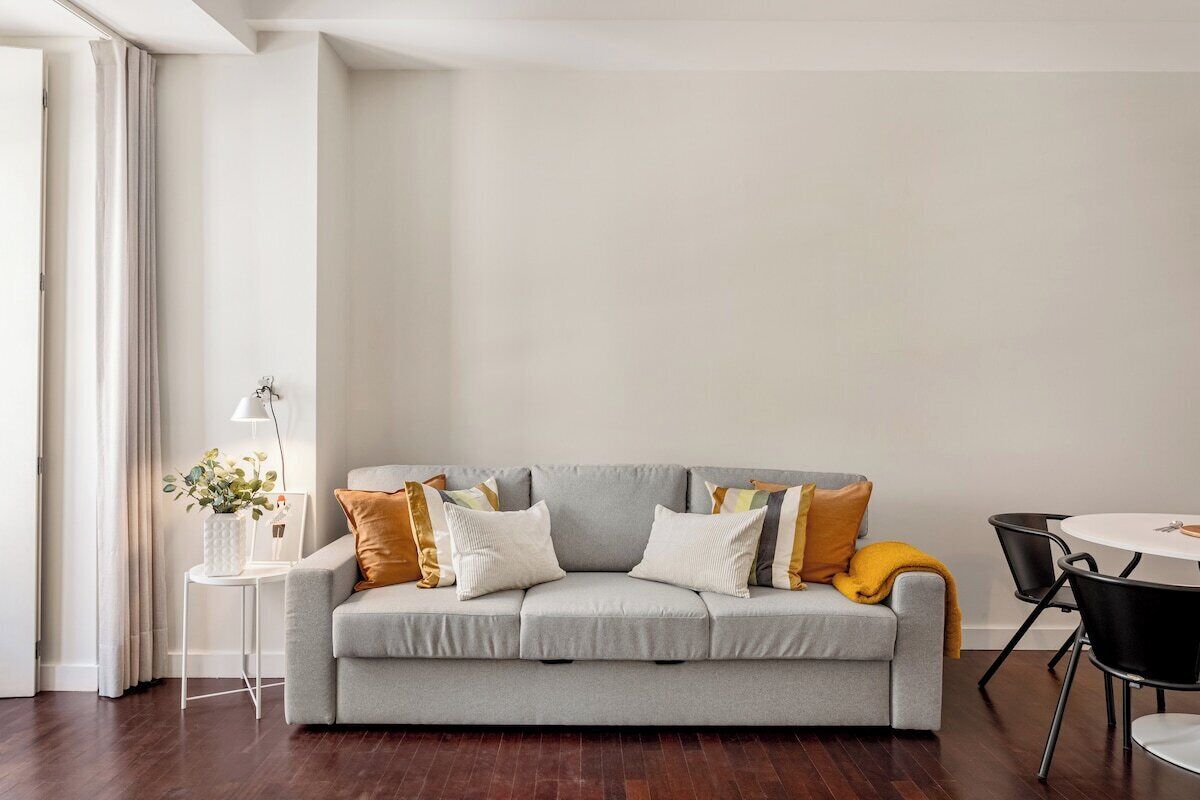 Uma sala de estar moderna com um sofá cinzento, uma mesinha de apoio com abajur e uma mesa de jantar redonda com cadeiras.