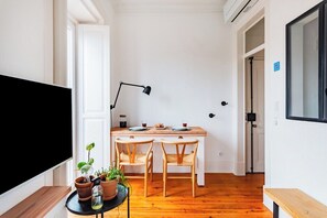 Ein kleines Zimmer mit einem Holztisch, zwei Stühlen, einer schwarzen Stehlampe und einem Fernseher links.